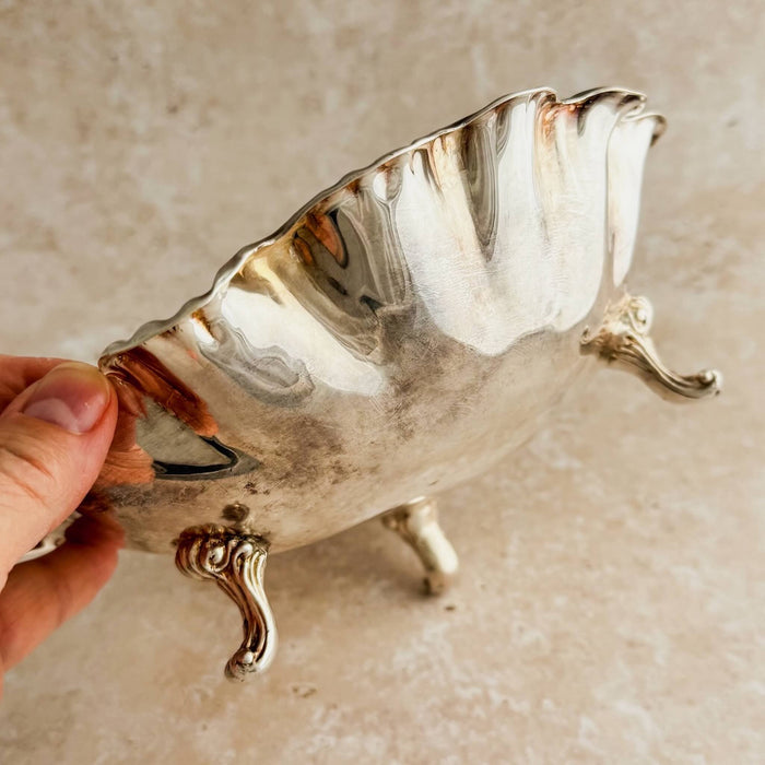 Side view of vintage silverplate footed bowl showing three ornate scroll feet and ruffled swirl rim, held to show scale and exterior patina