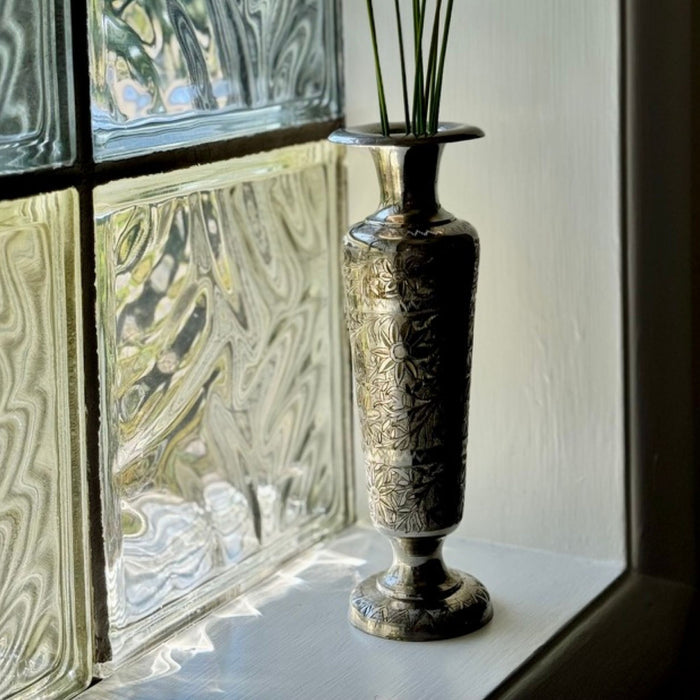 Vintage engraved metal bud vase holding slender green stems on a windowsill beside a textured glass block window.