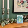 Vintage silver and lucite candle holders with taper candles styled on a mantel with garland, seasonal artwork, and collected decor