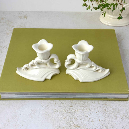 Pair of off-white Art Deco ceramic candle holders with flower-shaped cups, scrolling openwork, and leaf-form bases styled on a olive green book