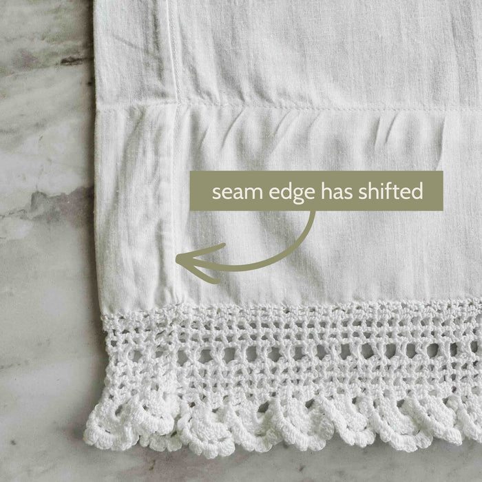 Close-up of one white pillowcase showing the side seam shifted inward above the crochet lace edge with an arrow and condition note