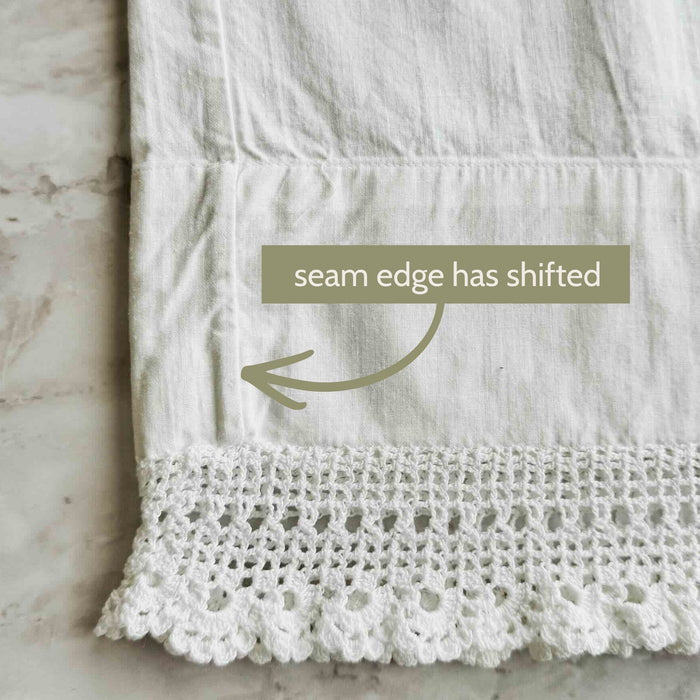 Close-up of the second white pillowcase showing the side seam shifted inward above the crochet lace edge with an arrow and condition note