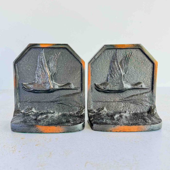 Pair of vintage cast iron bookends facing forward, each showing relief panel of a duck in low flight over water, dark finish with copper-toned highlights