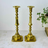 Pair of tall brass candlesticks with long decorative stems, hexagonal candle cups, and square bases with clipped corners styled with a trailing plant and book