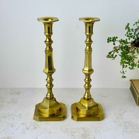 Pair of tall brass candlesticks with long decorative stems, hexagonal candle cups, and square bases with clipped corners styled with a trailing plant and book