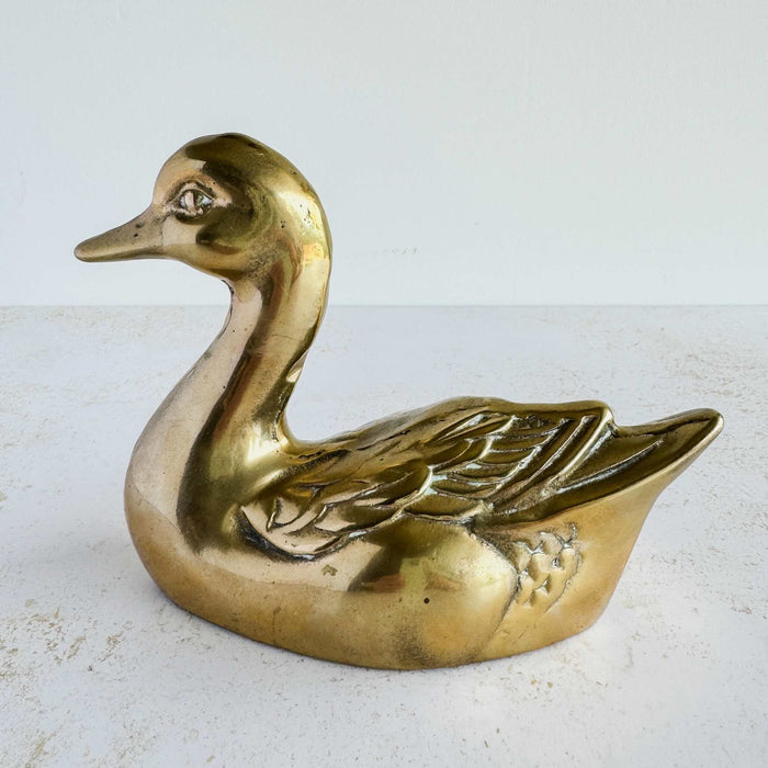 Vintage brass duck figurine shown from the side, with aged patina, raised wing details, and swimming pose