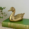 Vintage brass duck figurine with warm patina and raised wing detail, styled on a green antique book with trailing plant