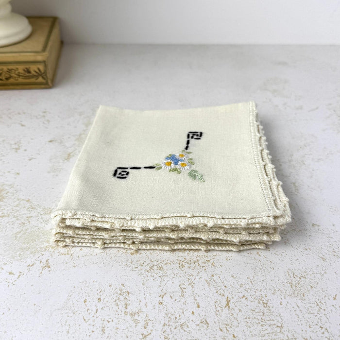 Stack of four folded vintage linen cocktail napkins with floral and black geometric hand embroidery and crochet edging