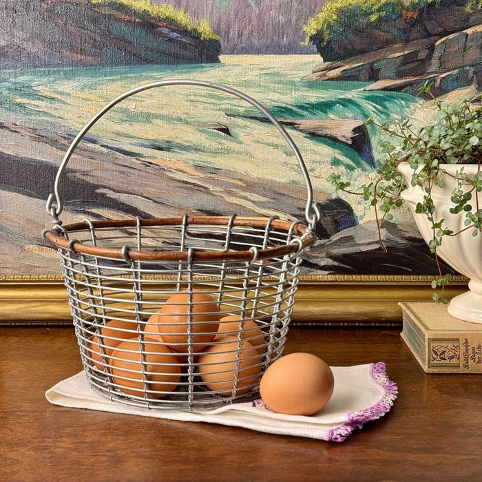 Vintage wire egg gathering basket with wood rim styled with brown eggs and a white embroidered kitchen towel with a framed landscape in the background