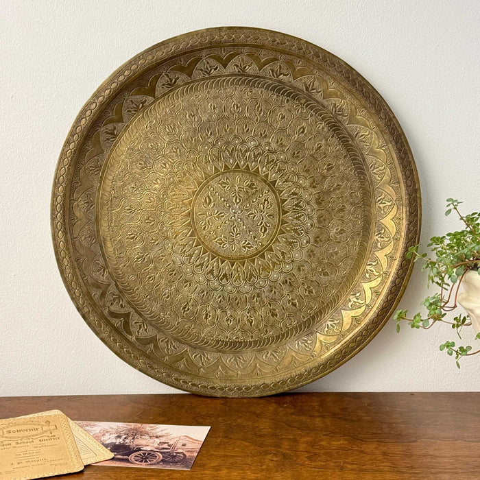 Vintage round brass tray with an intricate floral and geometric design, shown upright against a light wall with warm aged patina