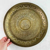 Vintage round brass tray held upright by hand, showing the complete decorative surface and aged patina