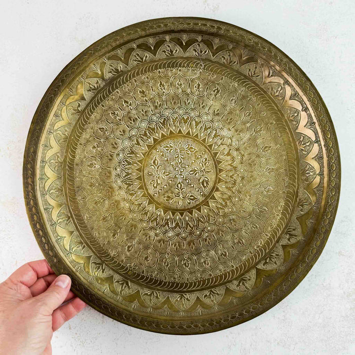 Vintage round brass tray held upright by hand, showing the complete decorative surface and aged patina