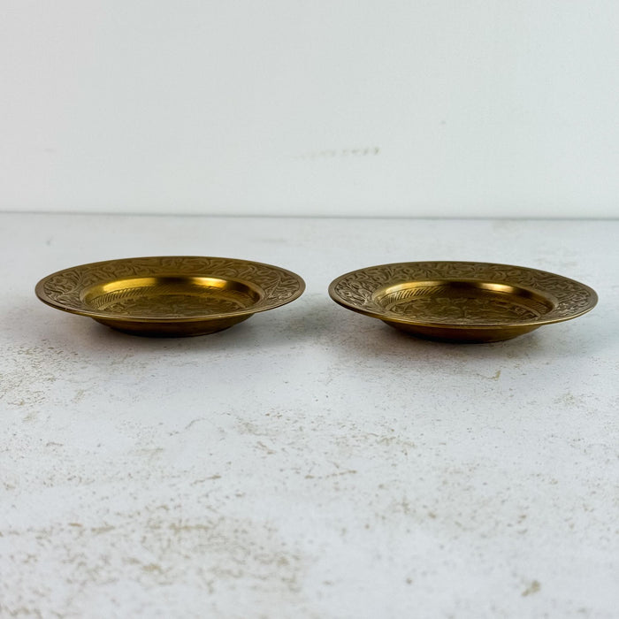 Pair of vintage engraved brass trinket dishes shown from the side on a light surface, revealing shallow bowl profile and wide engraved rim