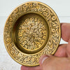Close-up of one vintage brass trinket dish held in hand, showing dense hand-engraved floral medallion at center and scrolling leaf border on wide rim