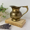 Vintage brass pitcher with rounded body, pedestal foot, curved handle, and aged patina, styled on an antique book with lavender and plant