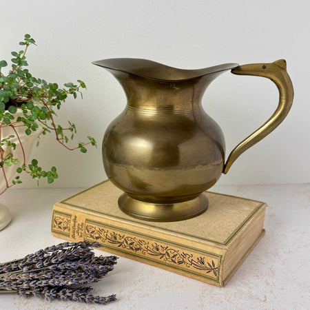 Vintage brass pitcher with rounded body, pedestal foot, curved handle, and aged patina, styled on an antique book with lavender and plant