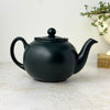 Back view of matte black teapot showing the curved spout, rounded body, loop handle, and ball-topped lid