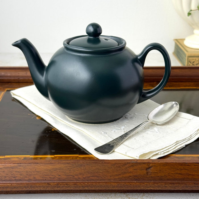 Matte black teapot styled on a folded white linen with a silver spoon on a dark wood tray