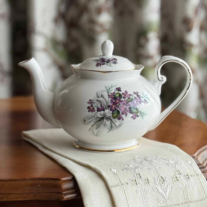 Vintage Royal Grafton English bone china purple violets teapot on antique table with linen tea towel and blurred floral drapery in background