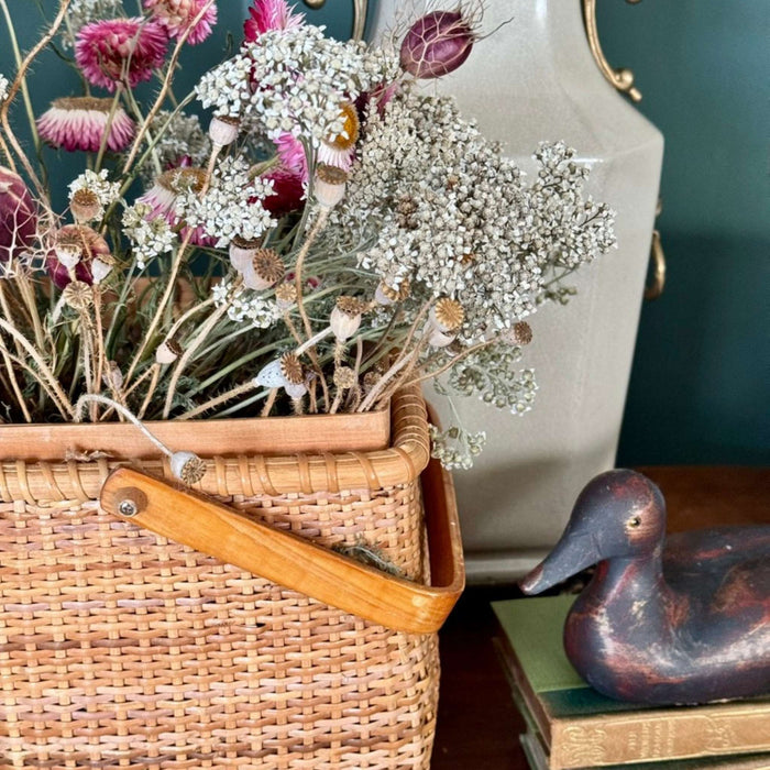 Vintage woven bamboo basket styled with dried flowers alongside a ceramic lamp, stacked books, and rustic duck decor