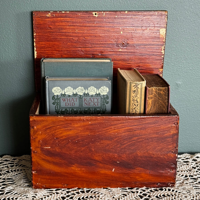 Antique primitive storage box with faux grain paint finish filled with vintage books against a muted green painted wall 
