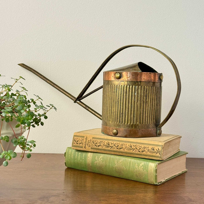 Vintage brass indoor watering can styled on green books with a long narrow spout, curved handle, ribbed body, copper bands, and aged patina