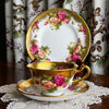 Royal Chelsea Golden Roses side plate standing behind matching teacup and saucer on a lace doily with blurred floral drapery in the background