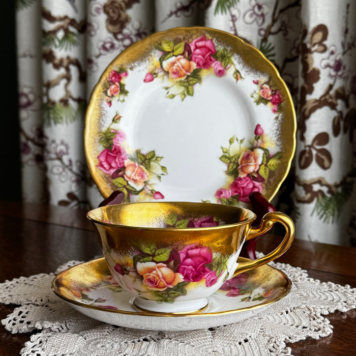 Royal Chelsea Golden Roses side plate standing behind matching teacup and saucer on a lace doily with blurred floral drapery in the background