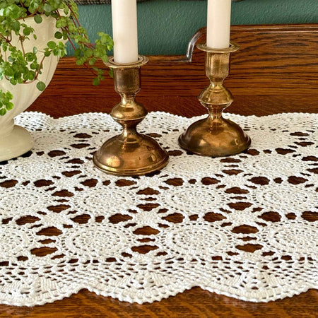 White crochet table runner styled across a dark wood surface with brass candlesticks and a white planter, showing the medallion pattern and scalloped edge.
