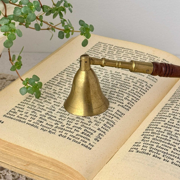 Vintage brass candle snuffer with bell-shaped end and turned wooden handle, styled on an open antique book with trailing plant