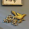 Vintage gold bird wall plaque with apple blossoms styled below framed botanical art on a gray-green wall