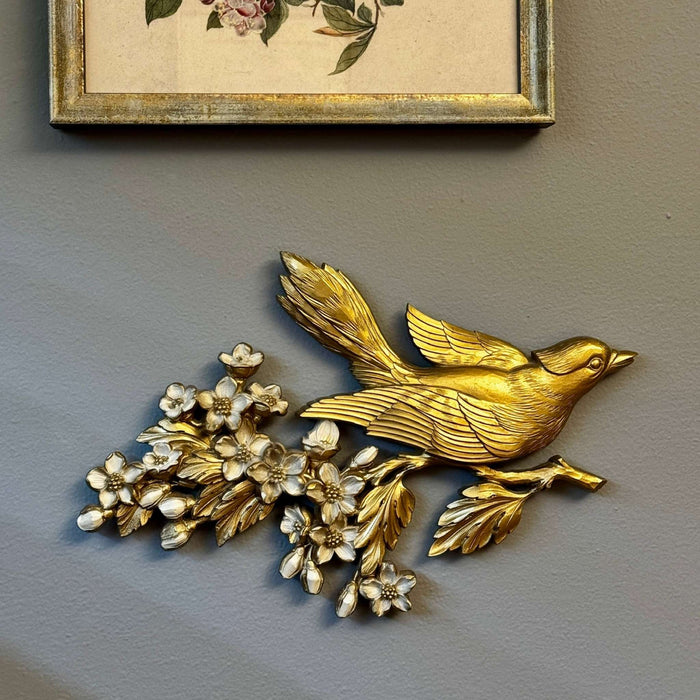 Vintage gold bird wall plaque with apple blossoms styled below framed botanical art on a gray-green wall
