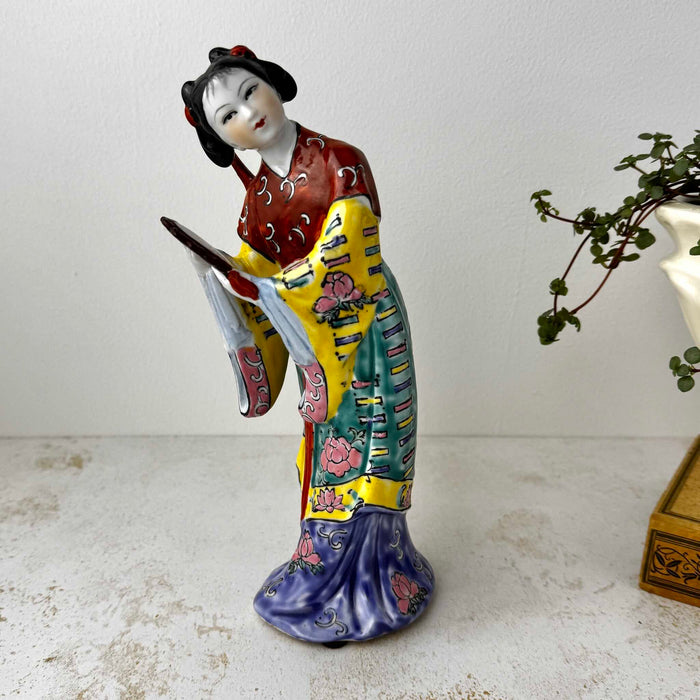 Vintage Chinese export porcelain figurine of a woman in a forward-inclined pose, holding a hand mirror, with robe painted in deep red, teal, yellow, and cobalt blue with pink peony motifs