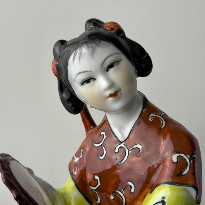 Close-up of hand-painted face on vintage Chinese porcelain woman figurine showing fine brushwork, arched brows, downcast eyes, and black sculpted hair
