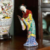 Vintage Chinese export porcelain figurine of a woman holding a hand mirror wearing a colorful robe, displayed on a wood table with a out-of-focus chinoiserie lamp and dark bookcase in the background