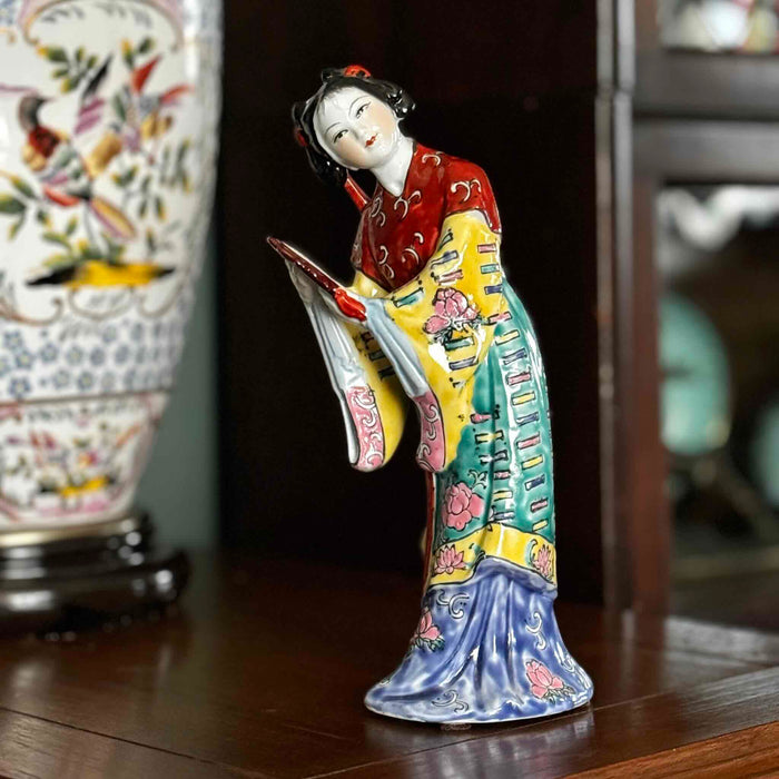 Vintage Chinese export porcelain figurine of a woman holding a hand mirror wearing a colorful robe, displayed on a wood table with a out-of-focus chinoiserie lamp and dark bookcase in the background