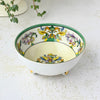 Small cream-colored porcelain bowl with a green rim, colorful bird and floral decoration inside, and three rounded feet with gold accents, displayed beside a trailing green plant.