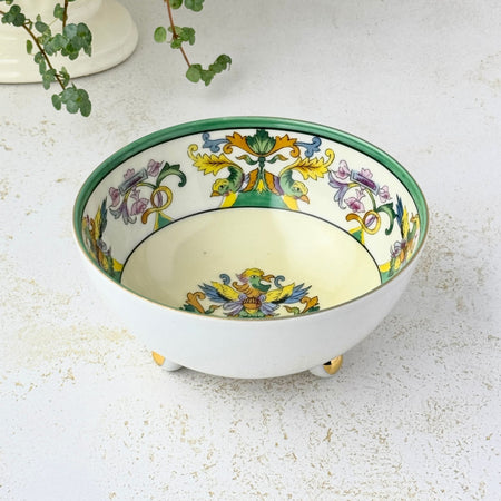 Small cream-colored porcelain bowl with a green rim, colorful bird and floral decoration inside, and three rounded feet with gold accents, displayed beside a trailing green plant.