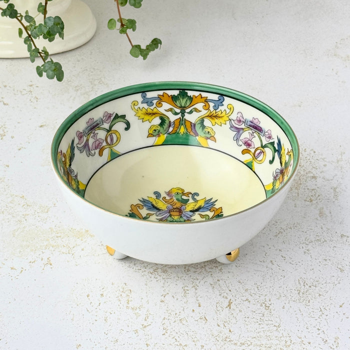 Small cream-colored porcelain bowl with a green rim, colorful bird and floral decoration inside, and three rounded feet with gold accents, displayed beside a trailing green plant.