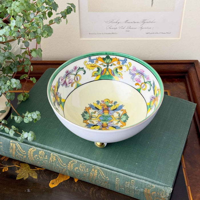 Vintage porcelain bowl resting on a green clothbound book, with its colorful interior decoration, green rim, and gold-accented feet visible beside a plant and framed botanical print.