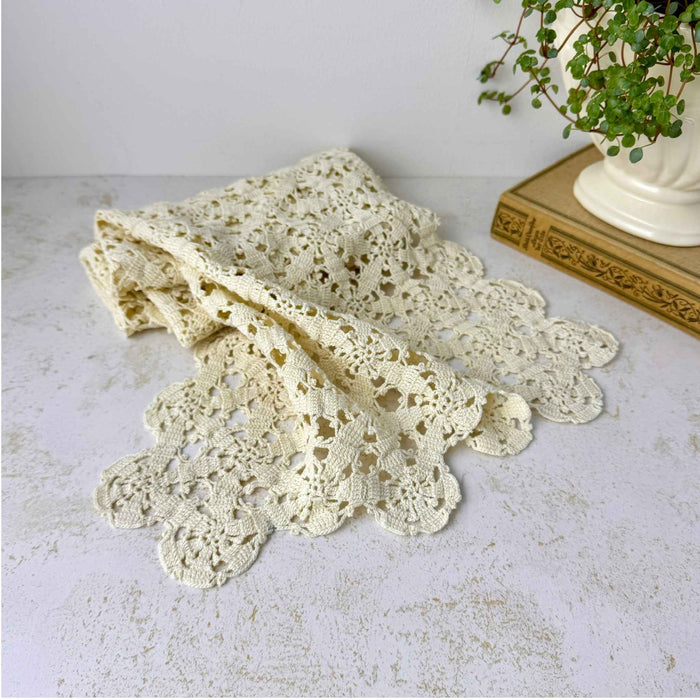 Folded ecru crochet table runner showing repeating floral rosettes, openwork pattern, looped details, and scalloped edge beside a potted plant and book.