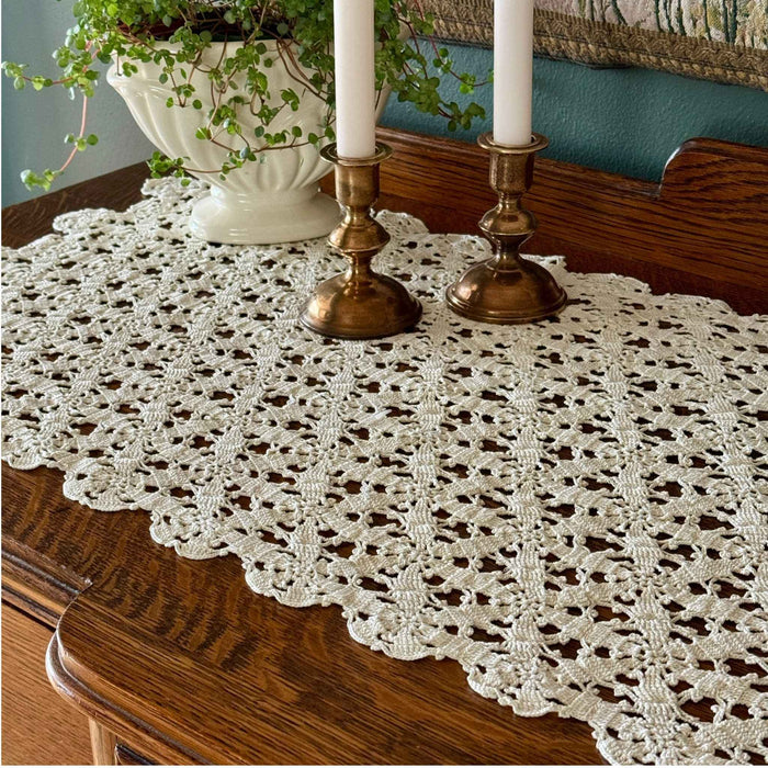 Ecru crochet table runner spread across a dark wood sideboard and styled beneath a white planter and two brass candlesticks.