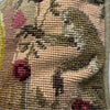 Detail of the wool needlepoint showing pink flowers, green foliage, and a small fanciful figure within the patterned background.