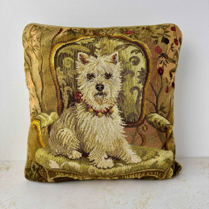 Square wool needlepoint pillow depicting a white Westie seated on an ornate armchair, surrounded by warm olive, gold, pink, plum, and tan details.