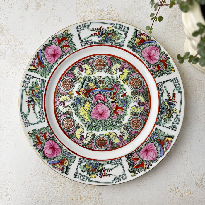 Vintage Chinese-style export porcelain plate with birds, peonies, butterflies, and longevity medallions in pink, turquoise green, and iron red, displayed on a white textured surface