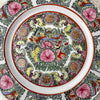 Close-up of center panel of vintage Chinese-style export porcelain plate with birds, peonies, butterflies, and longevity medallions in pink, turquoise green, and iron red, displayed on a white textured surface