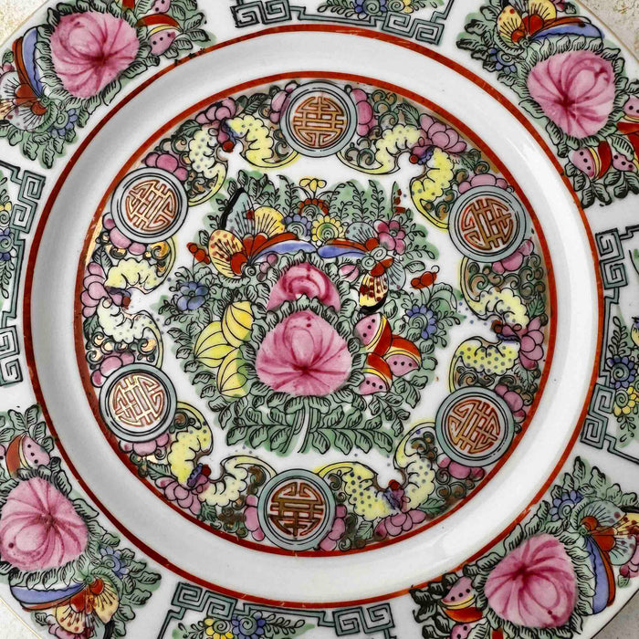Close-up of center panel of vintage Chinese-style export porcelain plate with birds, peonies, butterflies, and longevity medallions in pink, turquoise green, and iron red, displayed on a white textured surface