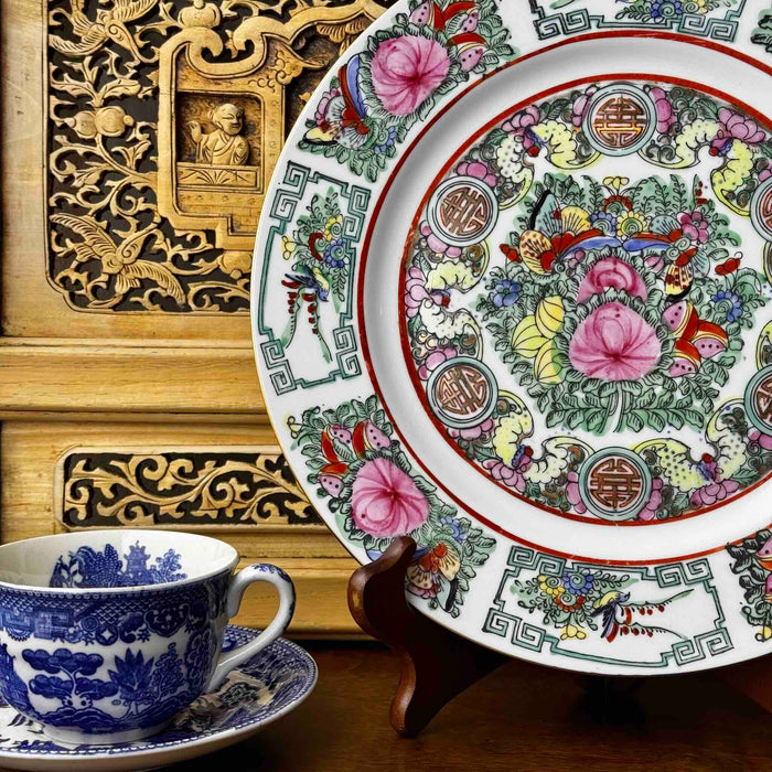 Vintage Chinese-style export porcelain plate with birds, peonies, butterflies, and longevity medallions in pink, turquoise green, and iron red, displayed on a plate stand against a Chinese carved wood wall panel and next to a Blue Willow teacup and saucer