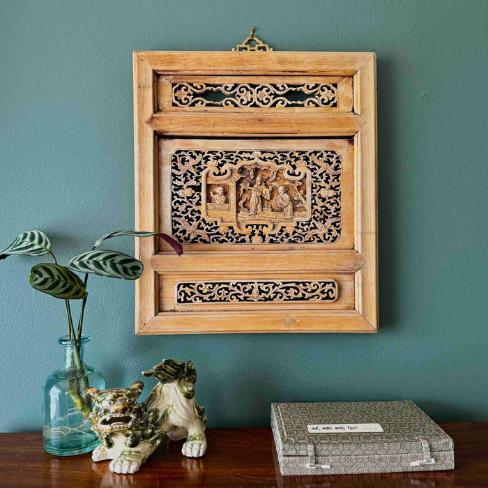 Vintage Chinese carved wood wall panel with openwork lattice surround and central relief scholar scene hanging on a blue-green wall with a plant, foo dog figurine, and calligraphy box on wood surface below