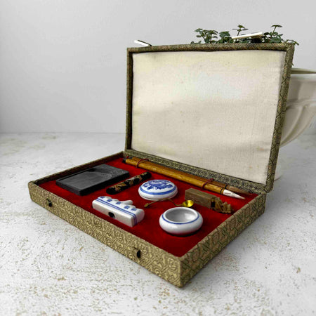 Vintage Chinese calligraphy set open in red-lined textile presentation box, showing ink stone, ink stick, two bamboo-handled brushes, porcelain seal paste container, brush rest, carved stone seal/chop, water dropper, and brass spoon in fitted recesses